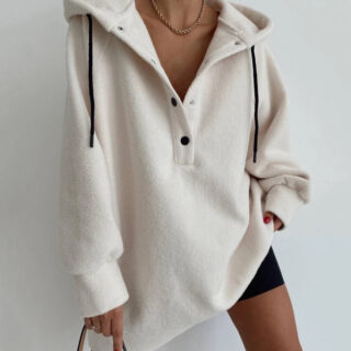 Photo représentant une jeune femme debout portant un pull polaire blanc à capuche long largement ouvert sur le devant la main gauche retenant un pan du pull sur un cycliste noir et la droite tenant l'anse d'un sac à main le tout sur un fond gris