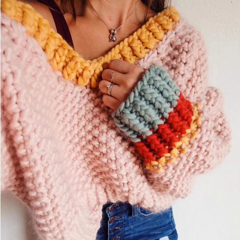 Photo montrant un pull tricoté en grosses mailles de laine rose à encolure jaune et côtes manches bleu ciel, rouge et jaune, porté par une femme main gauche repliée sur le coeur portant également un jean bleu foncé le tout sur un fond gris clair