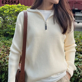 Photo d'une femme debout dans un jardin un sac marron en bandoulière sur l'épaule droite et portant un pull zippé beige à manches longues ouvert sur un tee-shirt blanc ras de cou et la main gauche dans la poche d'un pantalon beige foncé