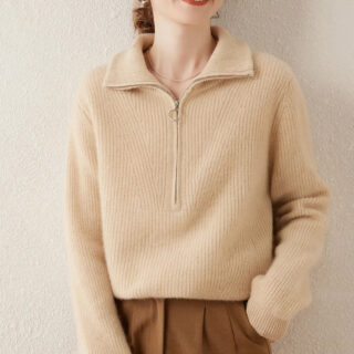 Photo d'une jeune femme debout main gauche dans la poche de son pantalon marron portant un pull zippé assez gros col ouvert beige à manches longues le tout sur un fond gris