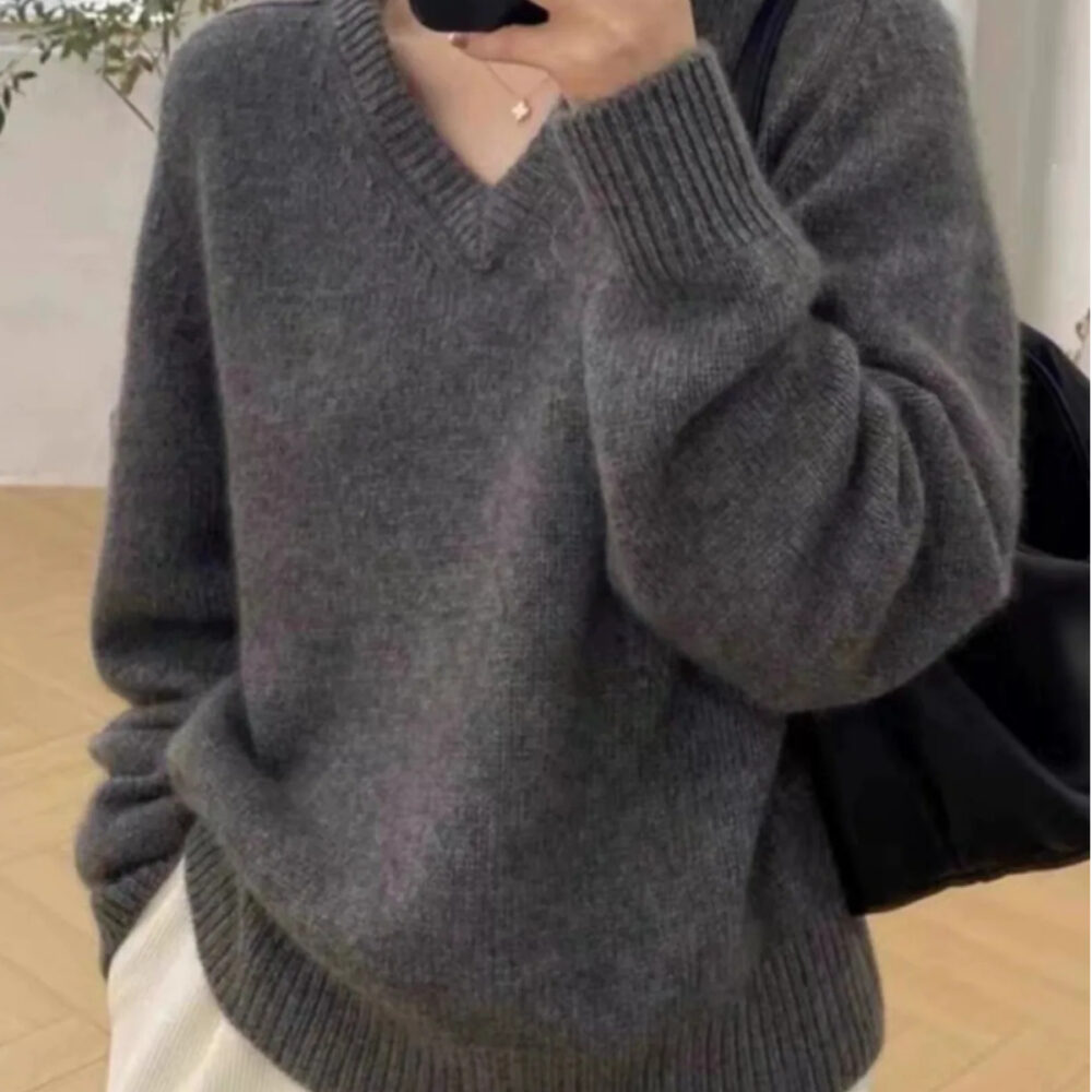 Photo représentant une jeune famme portant un pull gris col V à manches longues en laine cachemire sur un pantalon blanc avec main dans la poche un sac noir sur l'épaule gauche photo prise dans une pièce au sol beige et mur gris