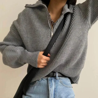 Photo d'une femme portant un pull camionneur gris ouvert sur un jean bleu ciel le bras gauche levé et la main droite sur la bandoulière d'un sac noir le tout sur fond gris