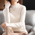 Pull col cheminé en maille chaude blanc porté par une femme avec un pantalon beige, un collier doré, asssise u un canapé gris.