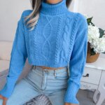 Photo d'une jeune femme assise devant une table de chevet garnie d'une plante fleurie blanche portant un jean taille haute bleu et un pull col roulé court torsadé bleu à manches longues