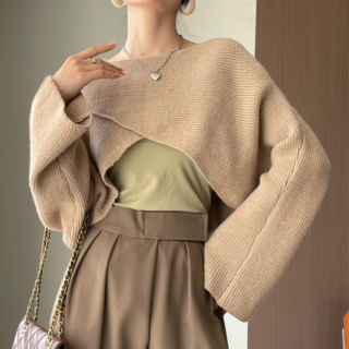 Photo d'une femme debout devant un mur gris et une porte marron foncé une main sur la hanche et l'autre vers le cou, portant un pantalon marron à pinces et un sous pull vert sur lequel un pull très court devant et croisé à larges manches finit le look avec un petit sac bandoulière sur le bras plié