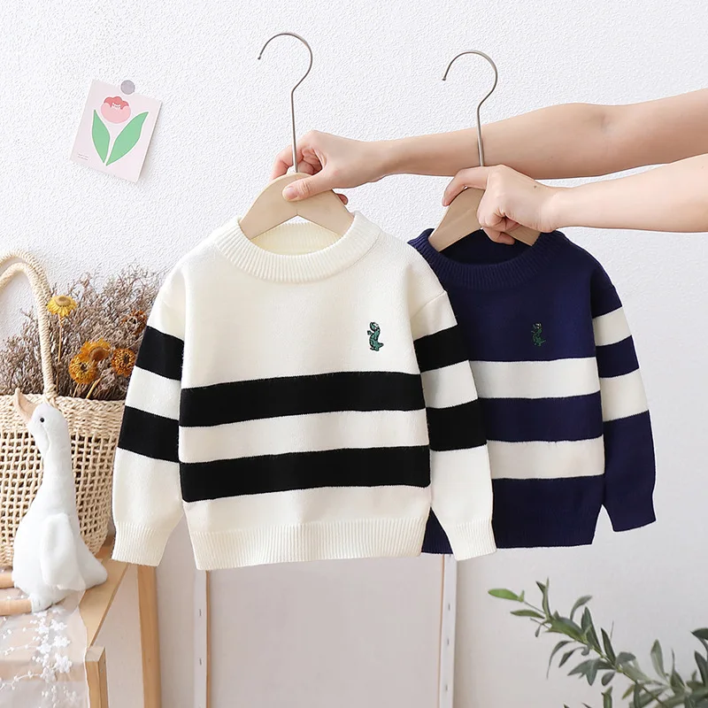 On voit une main qui tient deux pulls en tricot sur cintre. Il y a un pull blanc et un pull noir, rayé.