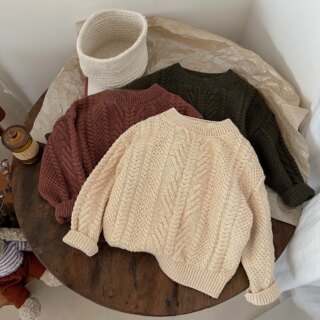 Sur une table ronde, dans un décor minimaliste, on voit trois pulls en tricot pour enfant.