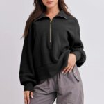 Photo d'une jeune femme portant un pull camionneur court noir sur un pantalon large gris foncé, la main gauche relevant légèrement le bas du pull le tout sur un fond gris clair