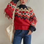 On voit une jeune femme qui porte un pull norvégien rouge, beige et noir, avec des touches de doré. Le pull est gai, avec des franges, lui donnant un air moderne.