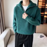 Photo d'un homme debout dans un salon devant un canapé et portant un pantalon foncé main droite dans la poche et un pull camionneur zippé vert la main gauche sur le zip ouvert