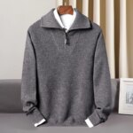 Photo d'un pull gris camionneur zippé à col moitié ouvert sur une chemise blanche elle-même sur un cintre en bois marron foncé au dessus d'un canapé gris devant un rideau beige