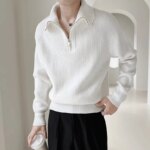 Photo d'un jeune homme debout portant un pantalon noir et un pull blanc camionneur zippé col ouvert le tout sur un fond gris clair flou