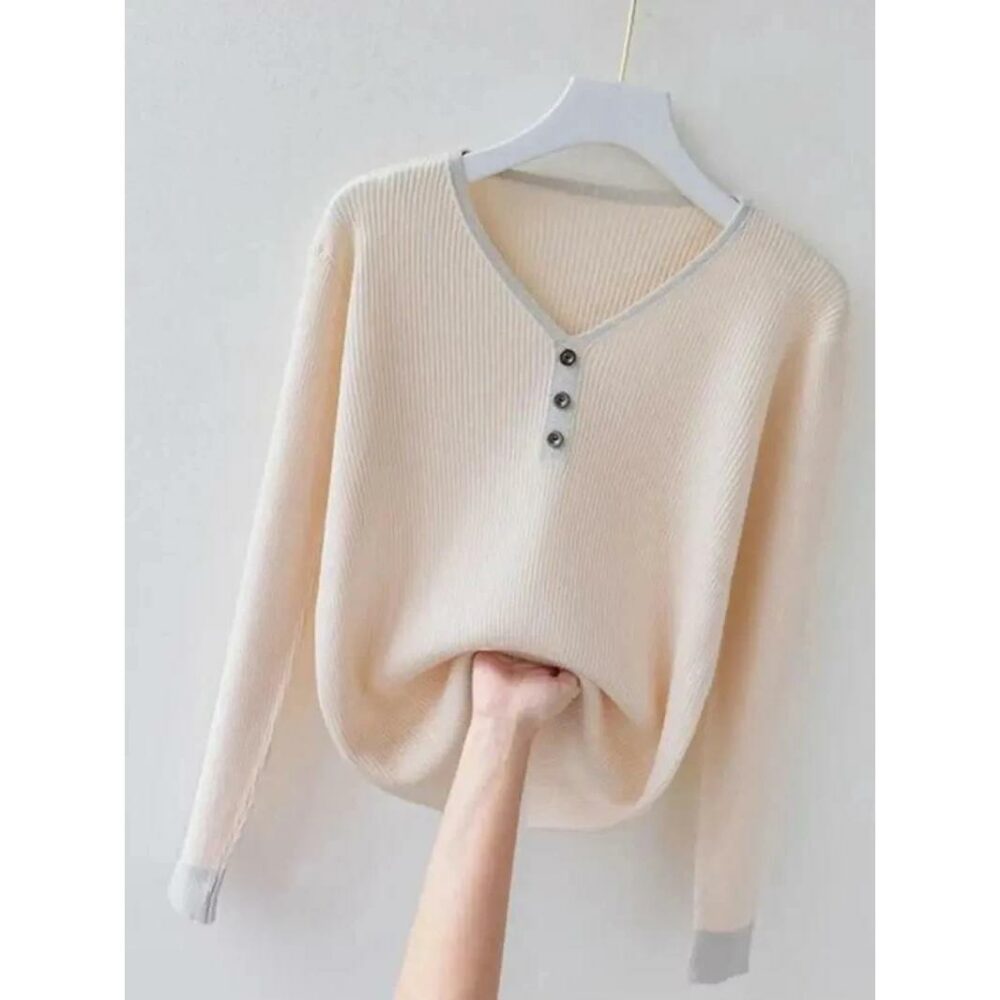 Photo d'un pull col V beige sur un cintre blanc en bois avec encolure 3 boutons, liseré autour du col et bas des manches d'un ton plus foncé, une main féminine remontant légèrement le bas du pull la tout sur un fond gris clair