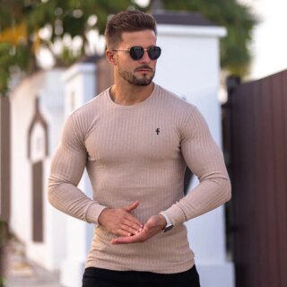 On voit un homme qui porte un sous-pull beige côtelé. Il est dehors avec des lunettes de soleil.