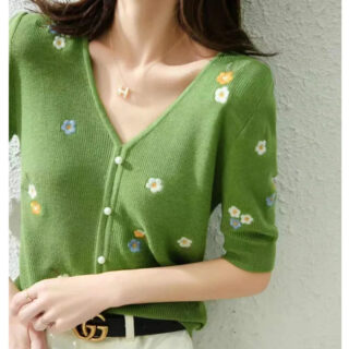 Photo d'une femme debout devant un mur gris le bras gauche replié et portant un pantalon beige à ceinture marron et boucle dorée et un pull vintage vert col V à petites fleurs brodées et fausse patte de boutonnage à boutons