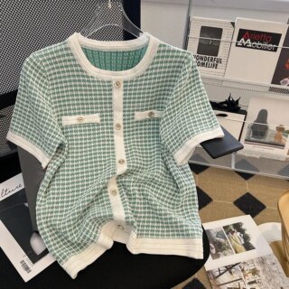 Photo d'un pull vintage femme vert d'eau et blanc à faux boutonnage devant et fausses poches , petits boutons argentés pendu sur un cintre transparent posé sur un siège noir avec des magazines au sol et sur un présentoir derrière à droite, un portefeuille sur une table basse transparente et un sol à carreaux style vintage