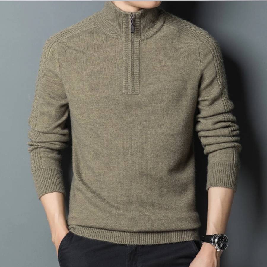 Photo d'un homme portant un pull zippé vert à fines torsades sur les épaules et les bras , manches légèrement relevéeset main droite dans la poche du pantalon noir, le tout sur un fond gris