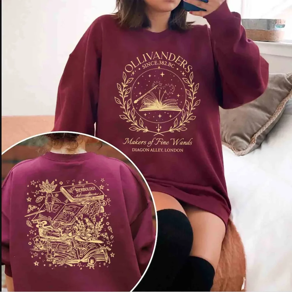 Photo d'une jeune femme assise sur un pouf rose et portant des guêtres noires et un pull bordeaux à col rond en sweat orné d'un imprimé doré représentant sur le devant un cercle en lauriers pour la boutique Ollivanders et au dos un encart carré montrant plusieurs détails significatifs de la saga sur une photo de dos enfermée dans un cercle