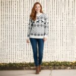 Femme caucasienne rousse en plain-pied portant un jean et un pull de Noël blanc à motifs imprimés nordique gris représentant des rennes et des flocons de neiges sur fond beiges.