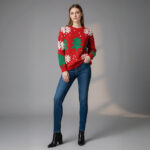 Jeune femme caucasienne en plain-pied posant et portant un pull de Noël rouge épais avec flocons de neige blanc et sapins de Noël tricotés sur fond gris.